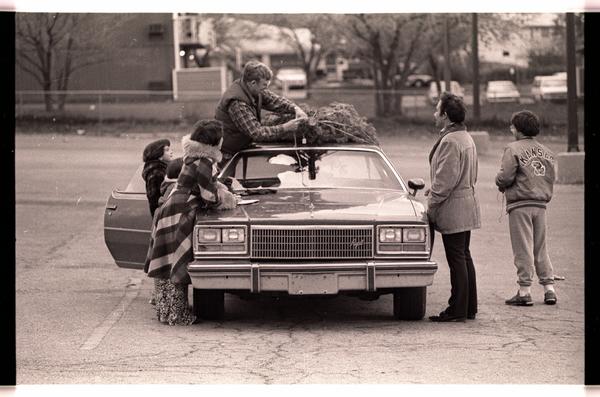 Sepia-toned photograph of five people standing around a car, one trying to tie a Christmas tree to the roof.