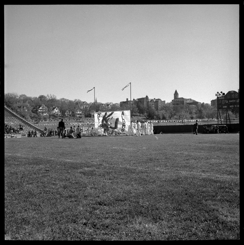 Photograph of KU football players running through a sign at the Homecoming game, 1958