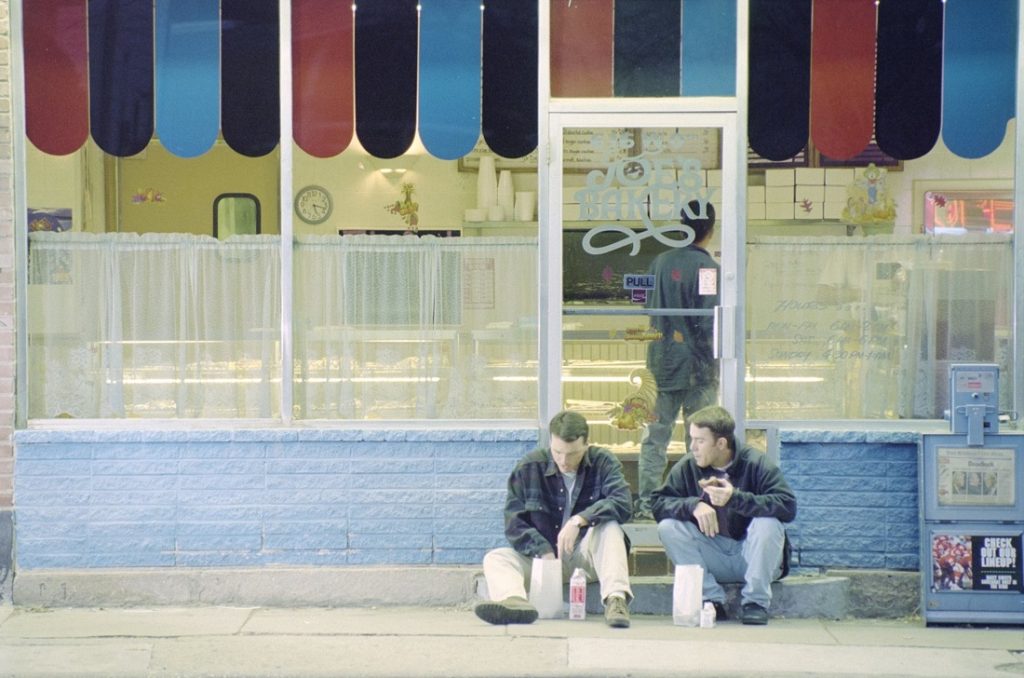 Photograph of two men sitting on the sidewalk outside Joe's Bakery, 1995