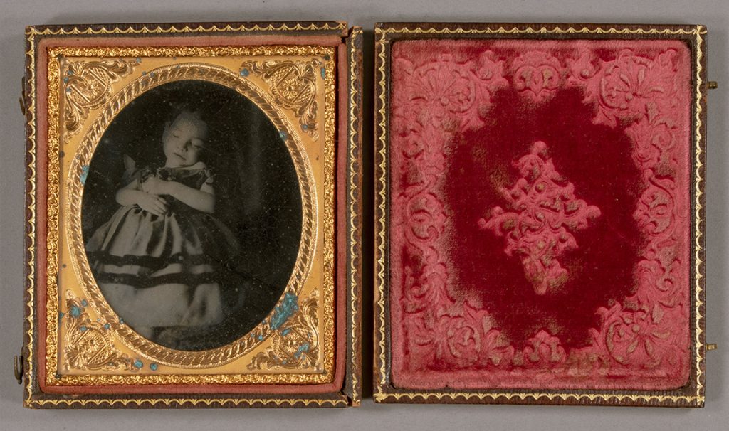 On the left is an oval sepia-toned photo with an embossed gold mat. The young girl in the photo is wearing a long dress; her eyes are closed and her hands are folded in front of her. On the right is a rectangle of embossed red velvet.