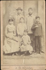 Cabinet card with identified students of Haskell Institute, photographer J. B. Shane of Lawrence. Students identified on the back as: 1. Geneva Roberts, Wichita (seated, far left); 2. Wiley Morgan, Seminole (standing, on left in back row); 3. Nellie Bates, Wichita (standing, center); 4. Nora Guy, Caddo (in front); 5. Peter Williams, Caddo (standing, on right in back row); 6. Richard Longhat, Caddo (standing, in dark uniform on far right).