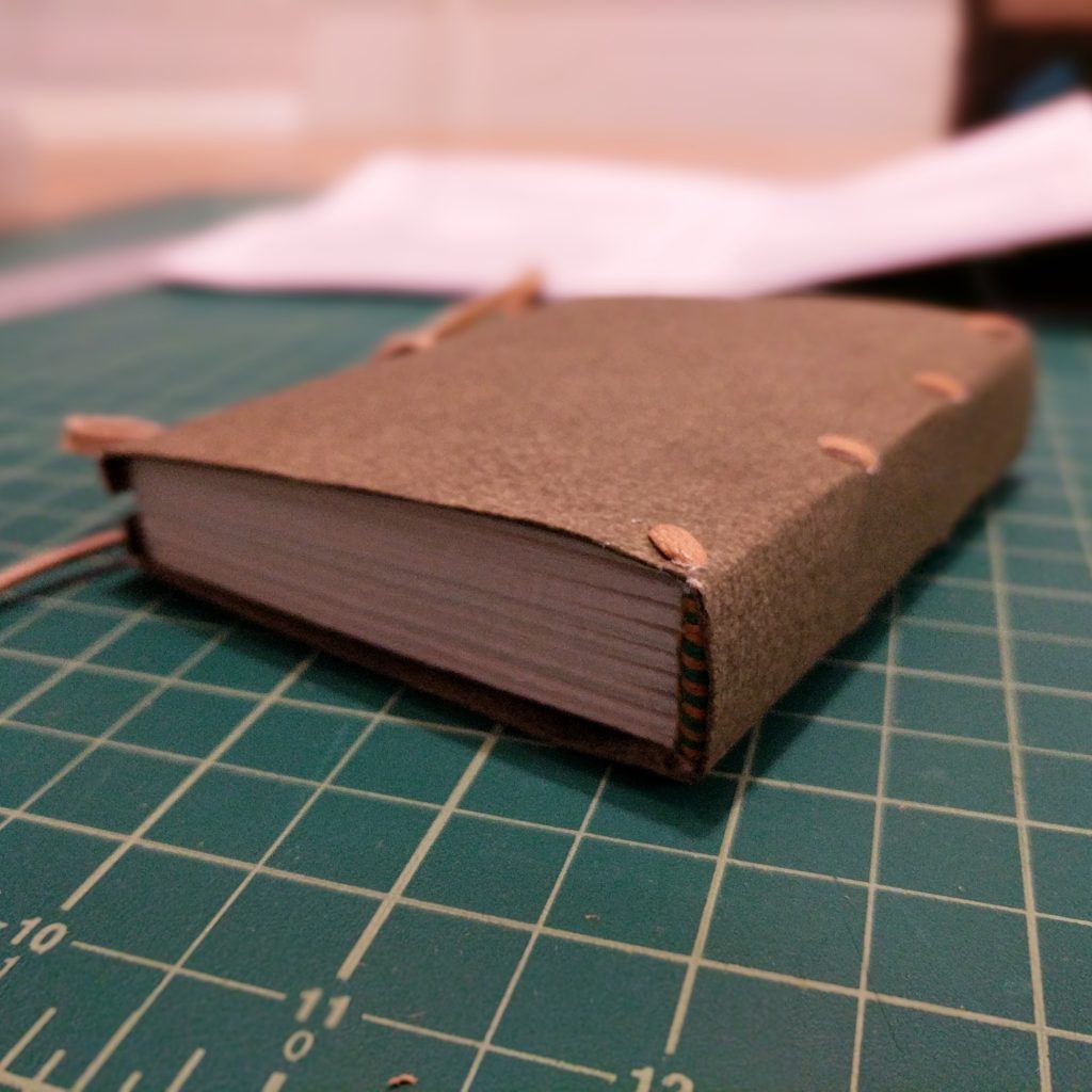 A small book lies on a cutting mat; the book is bound in the limp binding style, with a laced paper case, green and yellow endbands, and a fore-edge tie closure.