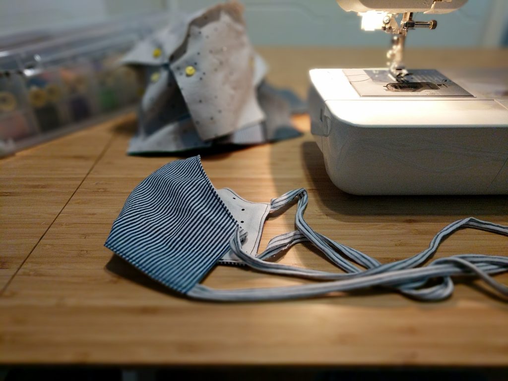 A handmade cloth face mask sits on a tabletop next to a sewing machine and other sewing supplies.