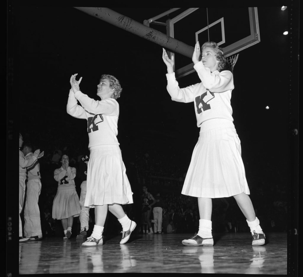 Photograph of KU cheerleaders clapping and cheering at a men's basketball game against Kansas State University, 1958-1959