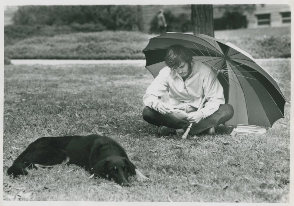 A black-and-white photograph of a male student sitting under an umbrella with a black dog.