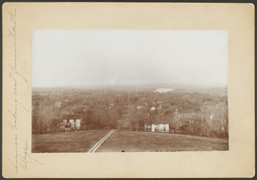 Black-and-white photograph of two people walking up Mount Oread on a pathway. Behind them, down the hill, are houses, trees, and unpaved streets.