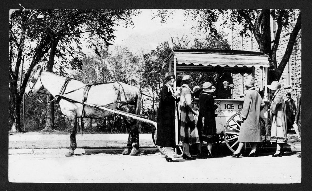 Black-and-white photograph of a horse-drawn wagon with an awning. Standing in front are five women wearing pumps, coats, and cloche hats.