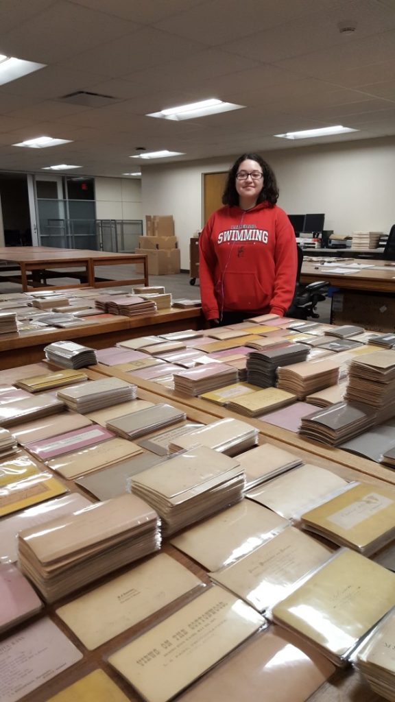 Young woman standing behind a large table covered with stacks of stereoviews, which are turned upside down.