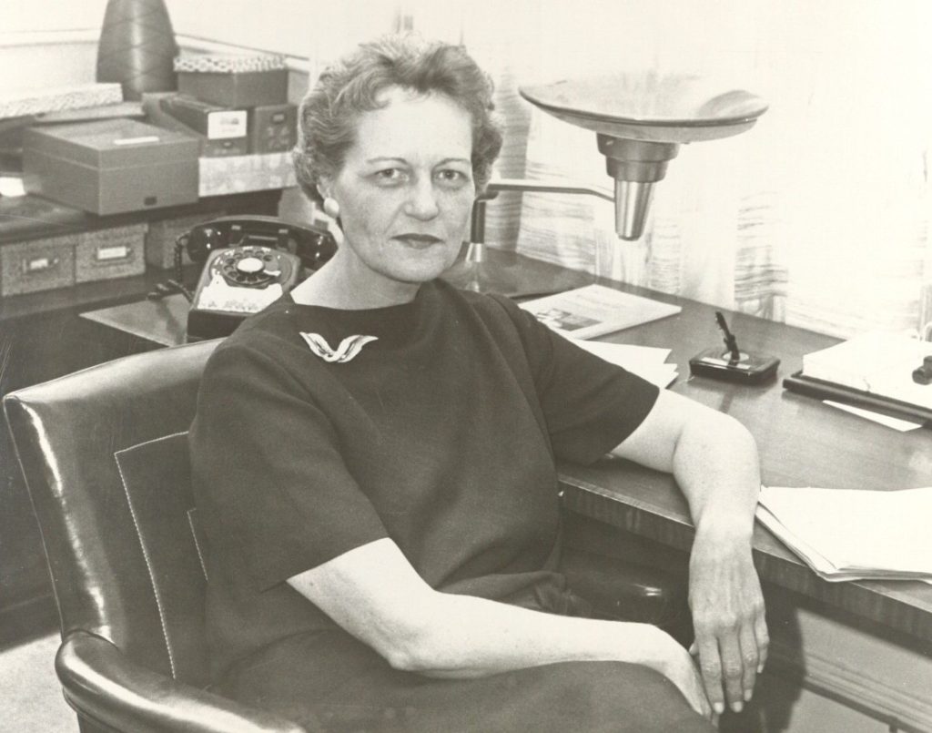 Black-and-white photograph of an older woman sitting at an angle. Her left elbow rests on a desk next to a black rotary phone, a lamp, and stacks of papers.
