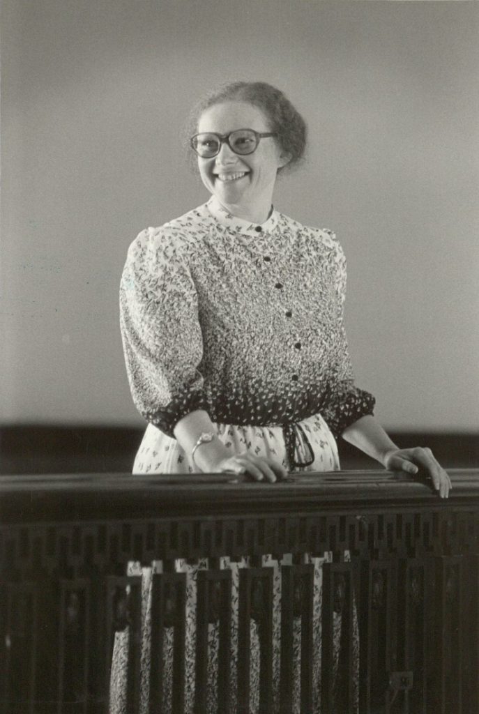 Black-and-white photograph of a woman in a patterned dress standing behind, with her hands on top of, a railing.