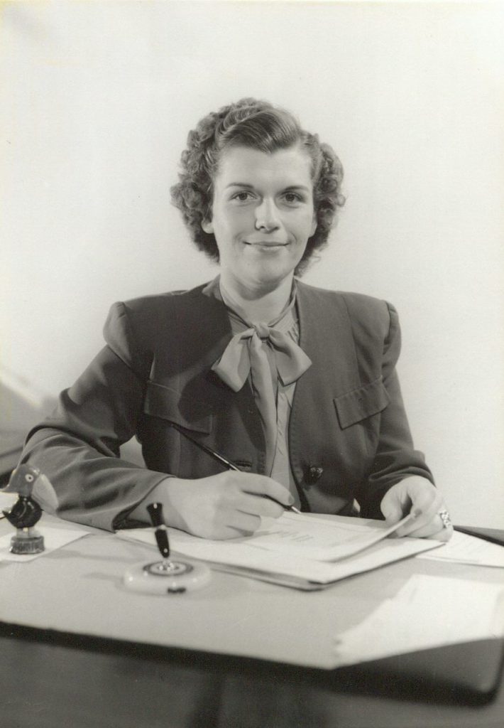 Black-and-white photograph of a young woman sitting at a table and writing. There is a Jayhawk figurine next to her right elbow.