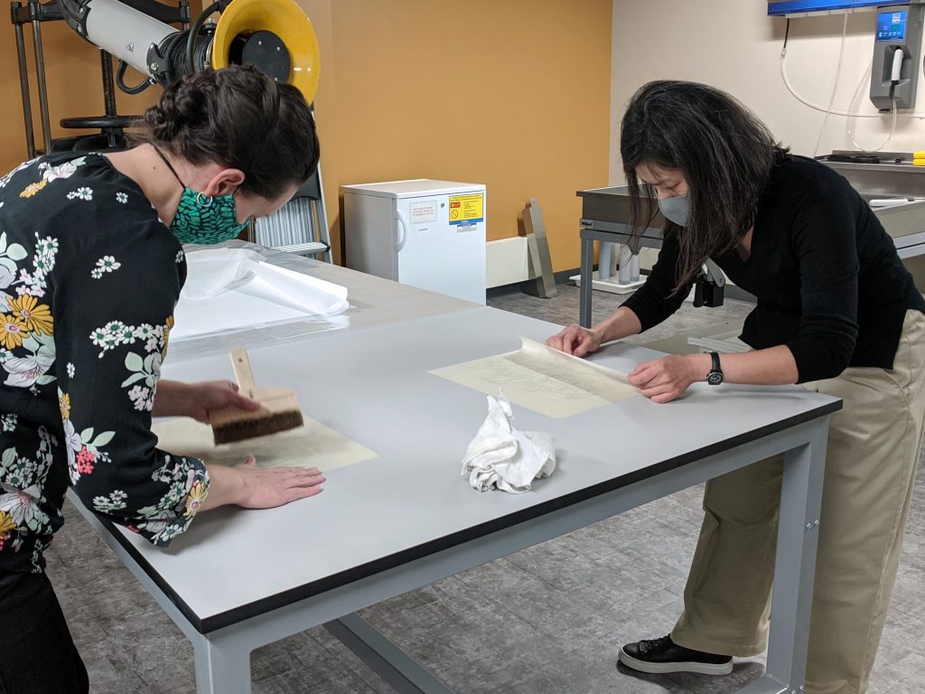 Mellon Initiative conservator Jacinta Johnson and visiting conservator Minah Song demonstrate friction drying, a technique for flattening delicate paper. Kenneth Spencer Research Library, The University of Kansas.