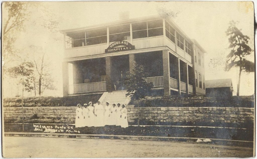 Black-and-white photograph of an enlarged two-story brick building with a front porch and a "Douglass Hospital" sign. A group of nurses stands in front.