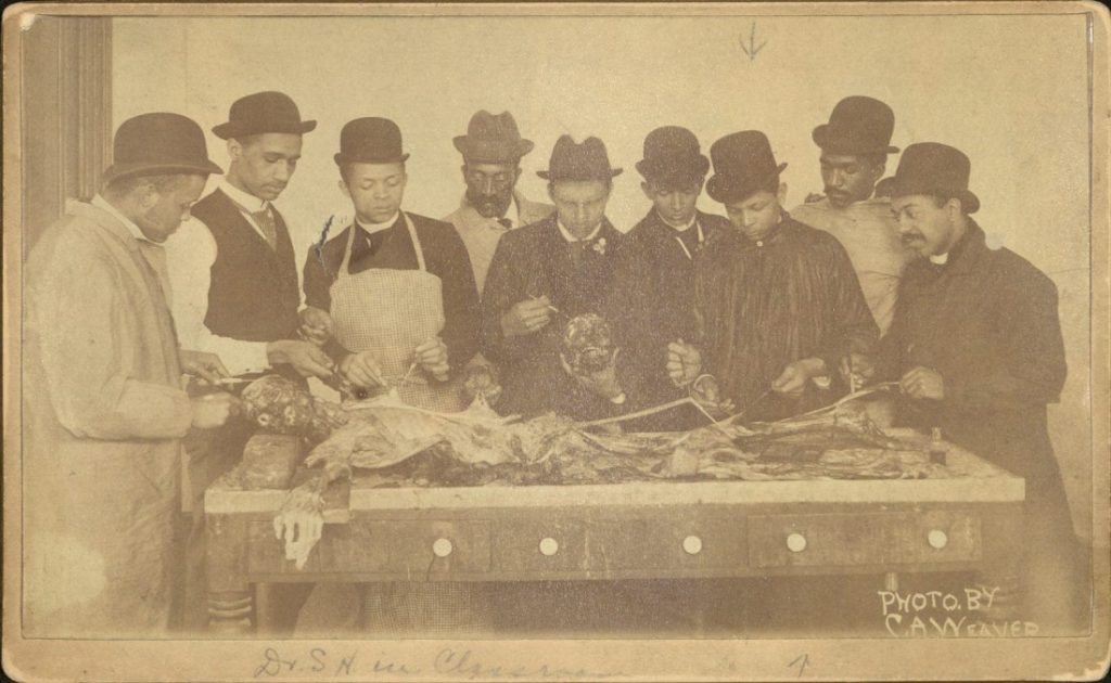 Sepia-toned photograph of men standing around a table. They are using medical implements to examine a human cadaver.