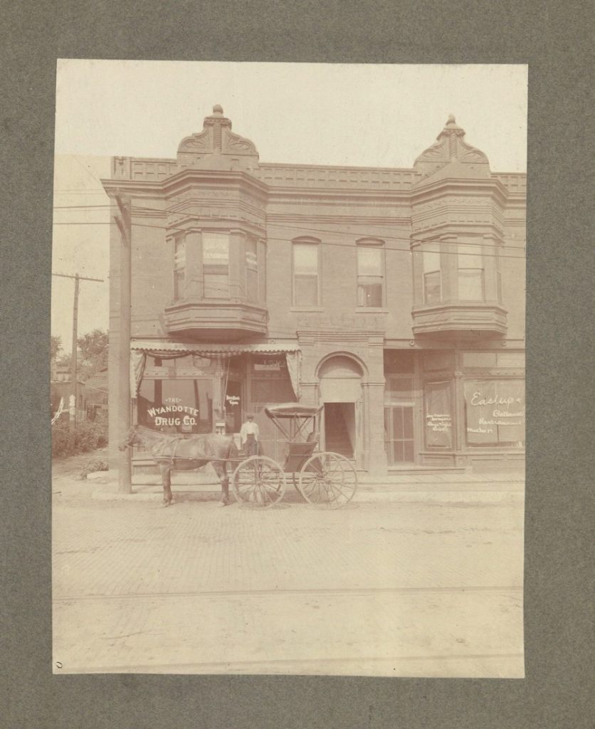 Black-and-white photograph of a two-story brick building with two second-floor bay windows. A man stands with a horse and buggy in front.