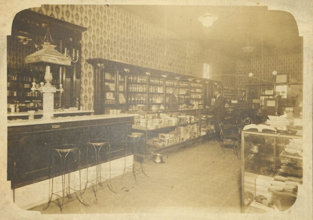 Sepia-toned photograph of two men, an employee and a customer, standing at glass display cases items. Full bookcases line the walls. On the left is a long counter with stools.