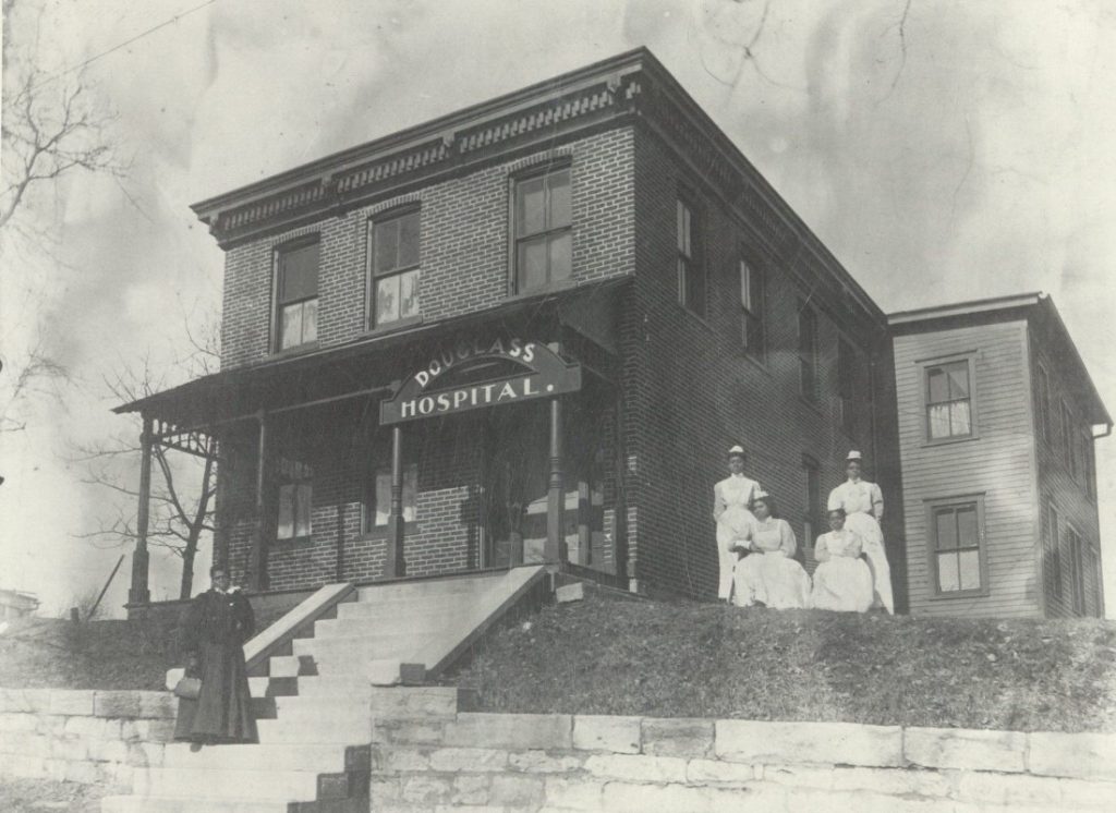 Black-and-white photograph of a two-story brick building with a front porch and a "Douglass Hospital" sign. Five women, four dressed as nurses, stand outside.