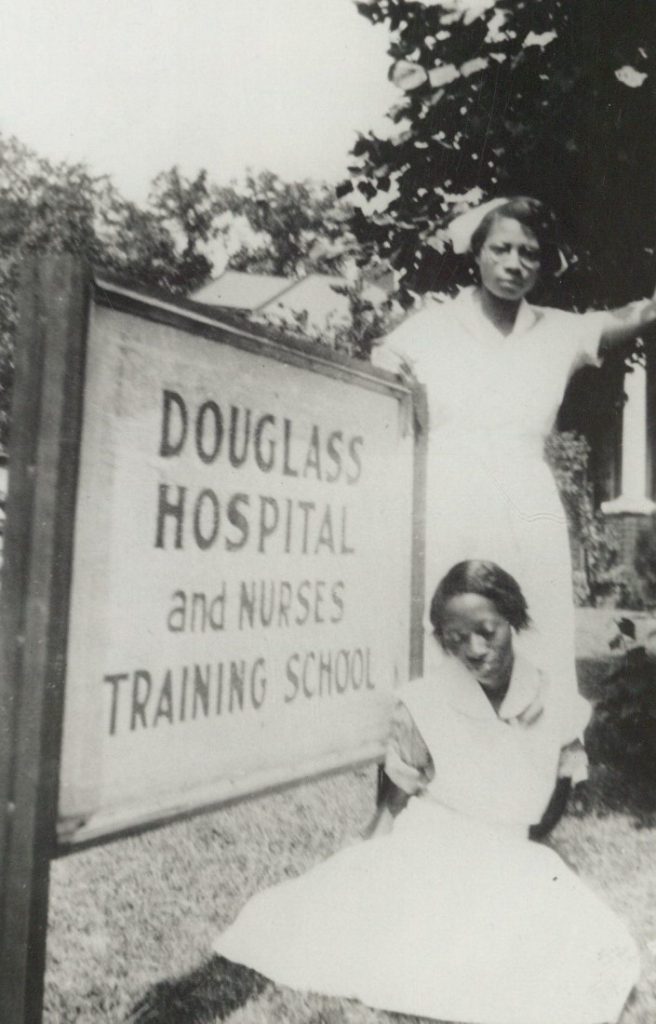 Black-and-white photograph of two students in nursing uniforms next to a sign that reads "Douglass Hospital and Nurses Training Program."
