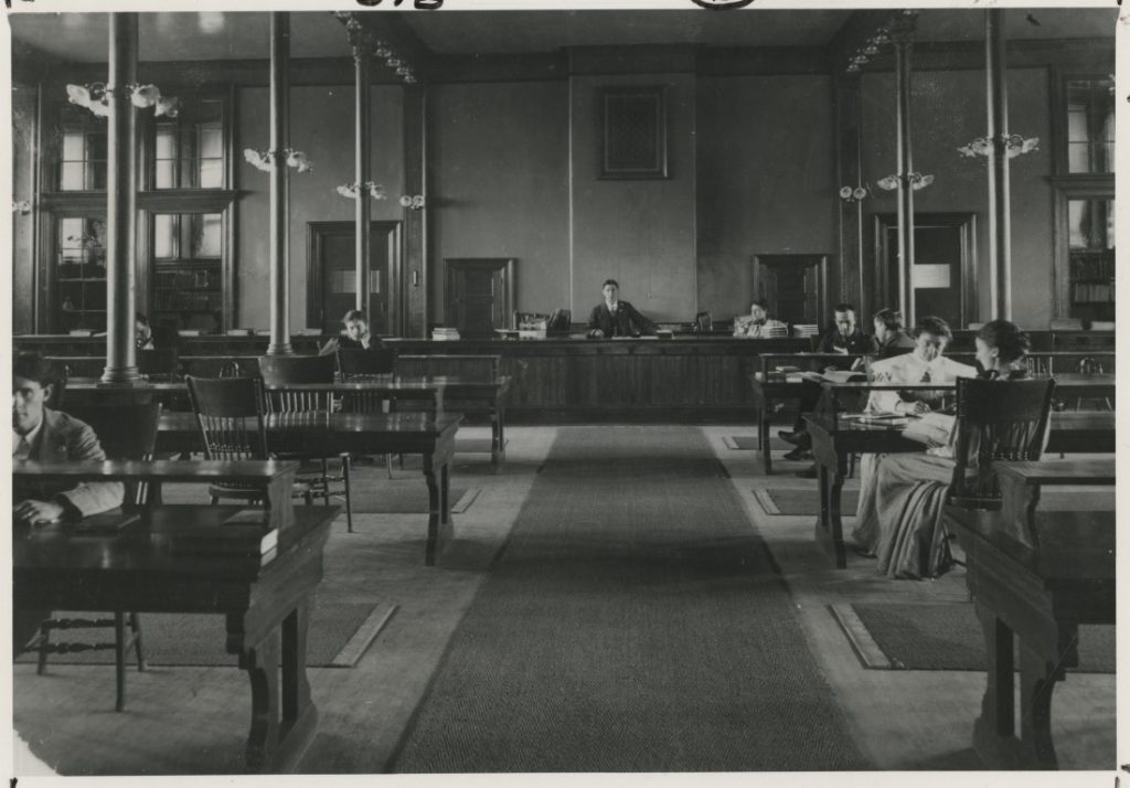 Black-and-white photograph of male and female students sitting and reading at wooden tables arranged in two rows with a cleared aisle in the middle.