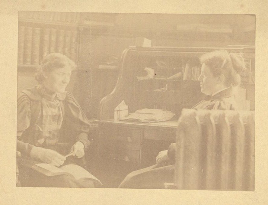 Sepia-toned photograph of two women sitting at a roll-top desk.