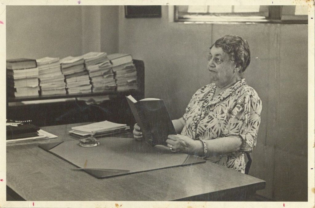 Black-and-white photograph of a woman sitting at a desk reading a book.