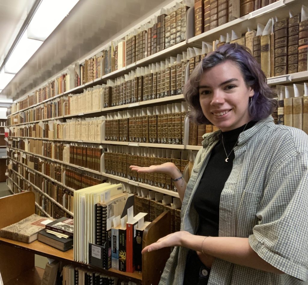 Young woman standing in front of floor-to-ceiling shelves of books, gesturing to a booktruck of materials.