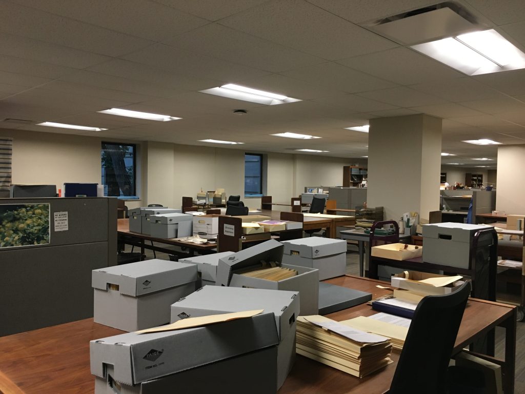 Photograph of a large open room. Large tables in the foreground are full of boxes and other library materials.