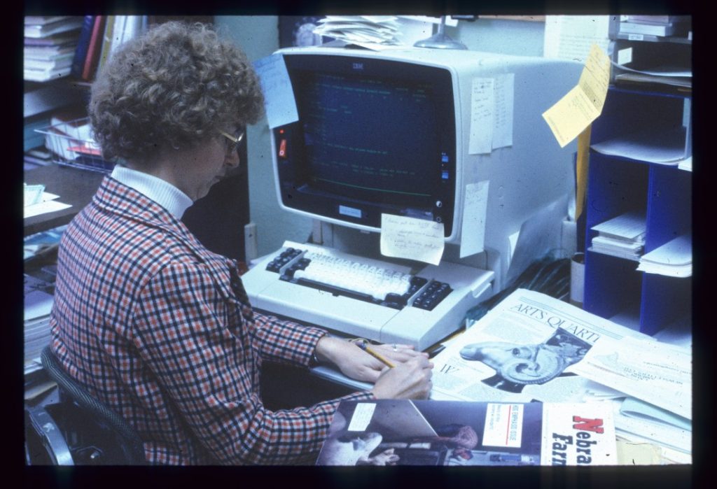 Color photograph of a woman sitting, with her back to the camera, facing a desktop computer. She is writing with a pencil, and there are library materials on her desk.