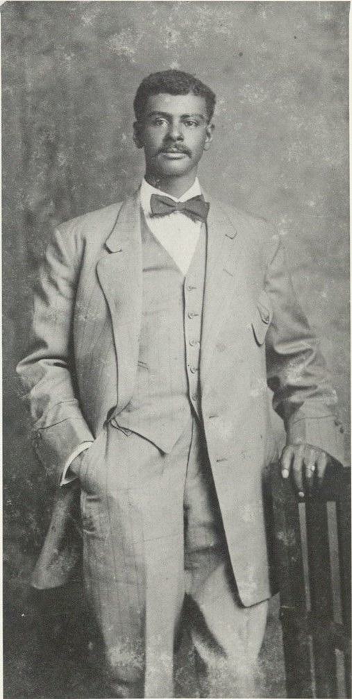 Black-and-white photograph of a young man wearing a suit. He is standing with one hand in his pocket and the other on the back of a chair.
