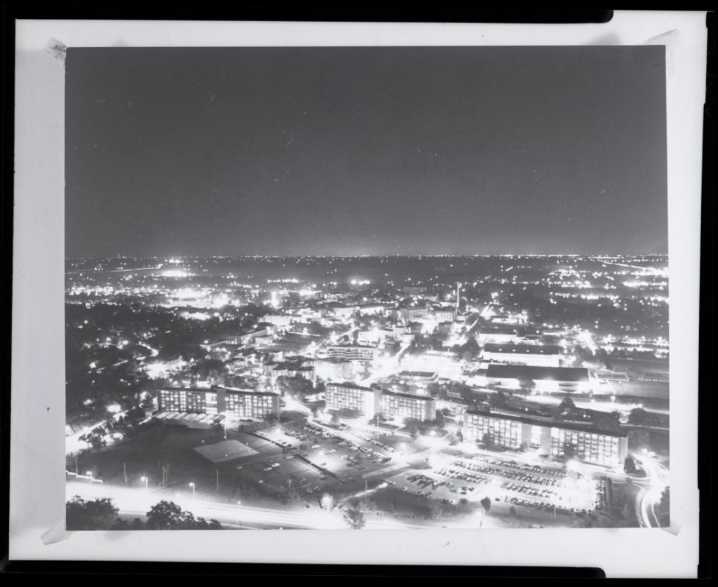 Black-and-white photograph of streets and buildings lit by bright lights.