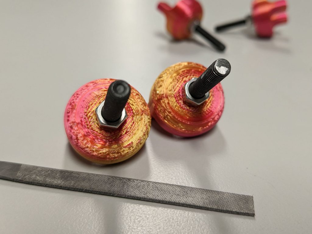 Two black metal bolts, each with a small silver hex nut and large red-and-yellow 3D printed nut on its end, sit on a table next to a small metal rasp. The end of the bolt on the left has been filed down smooth.