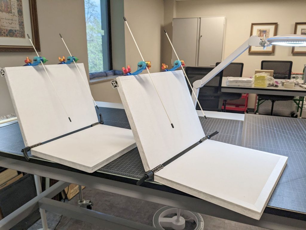 Two adjustable book cradles sit atop a workbench in a conservation lab.