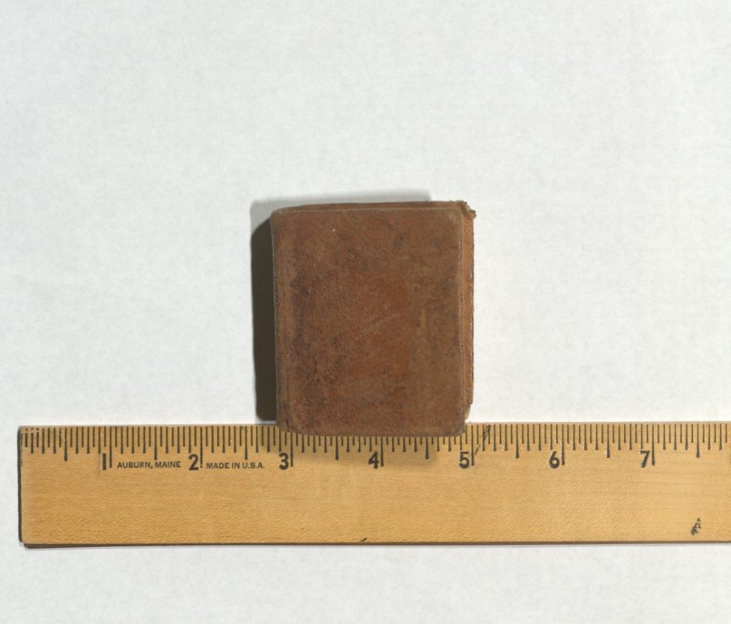 Book with a plain brown leather cover sitting above a ruler showing that the volume is about two inches wide.
