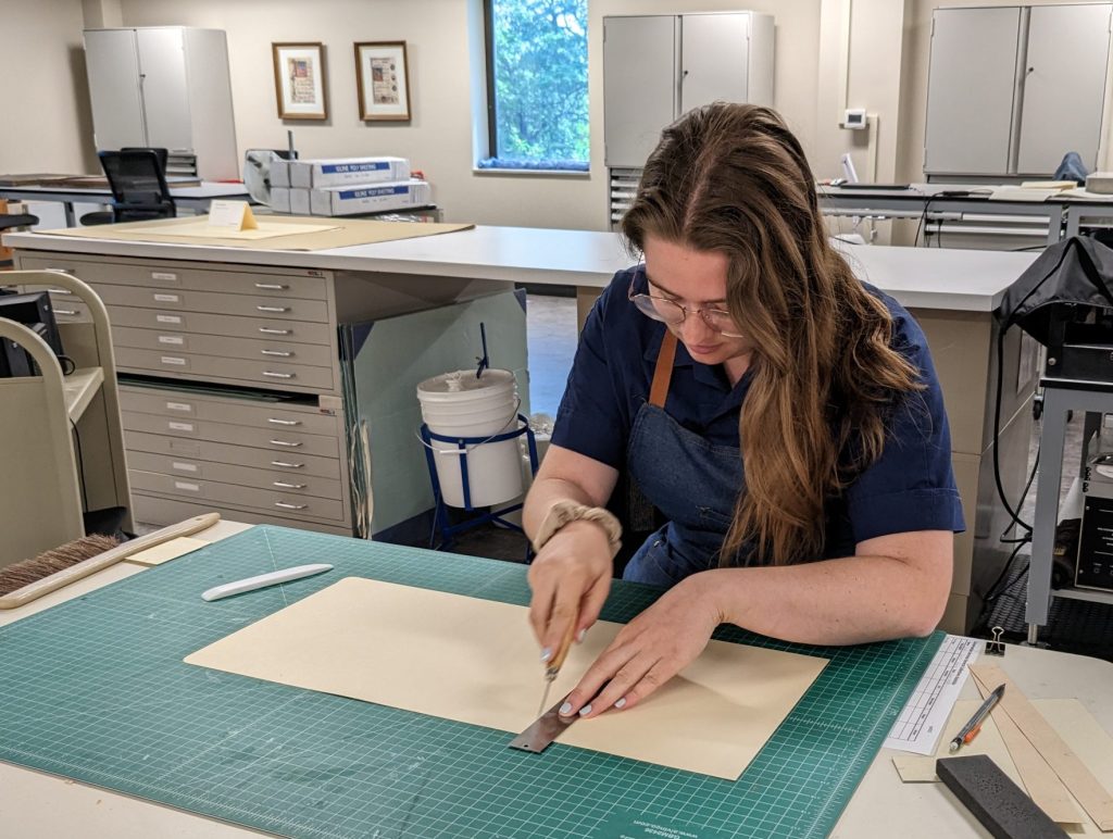 Conservation Services student assistant Sarah Jane Dahms sits at a workbench in the conservation lab while scoring a piece of board for a tuxedo box.