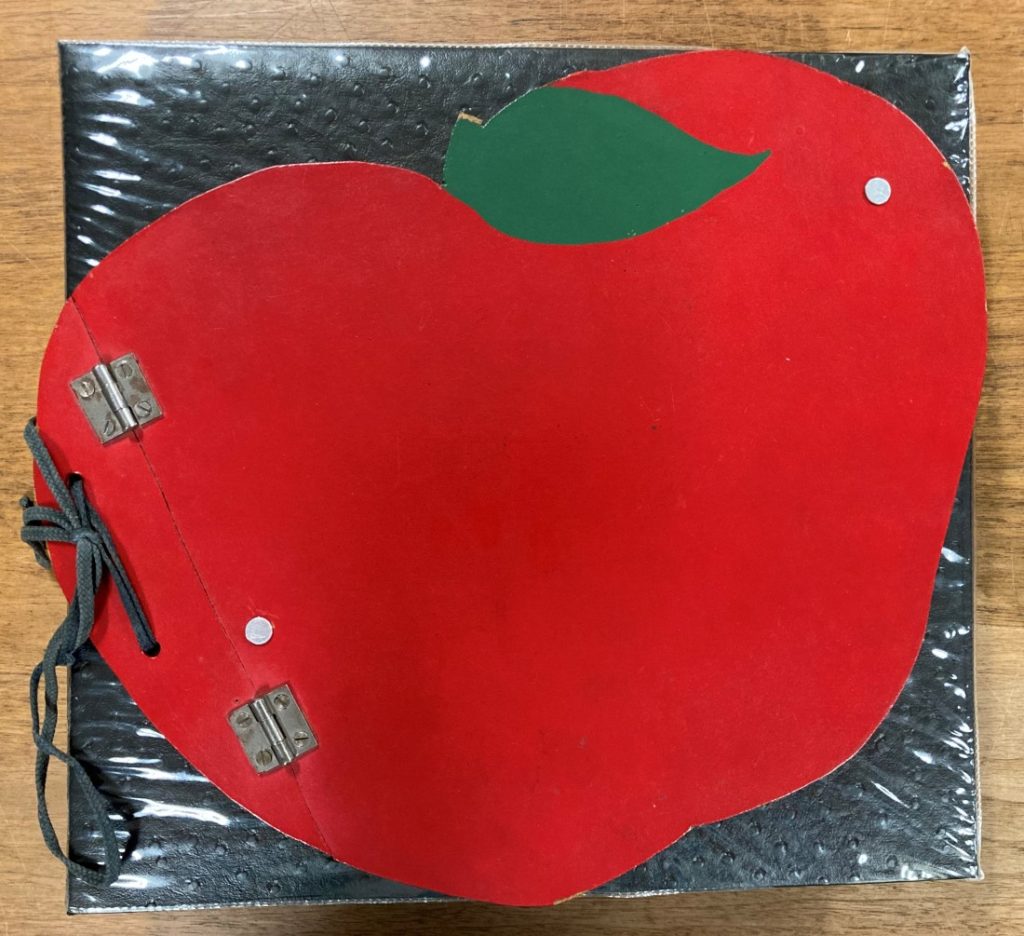 Wooden red apple on top of and affixed to a black scrapbook cover.