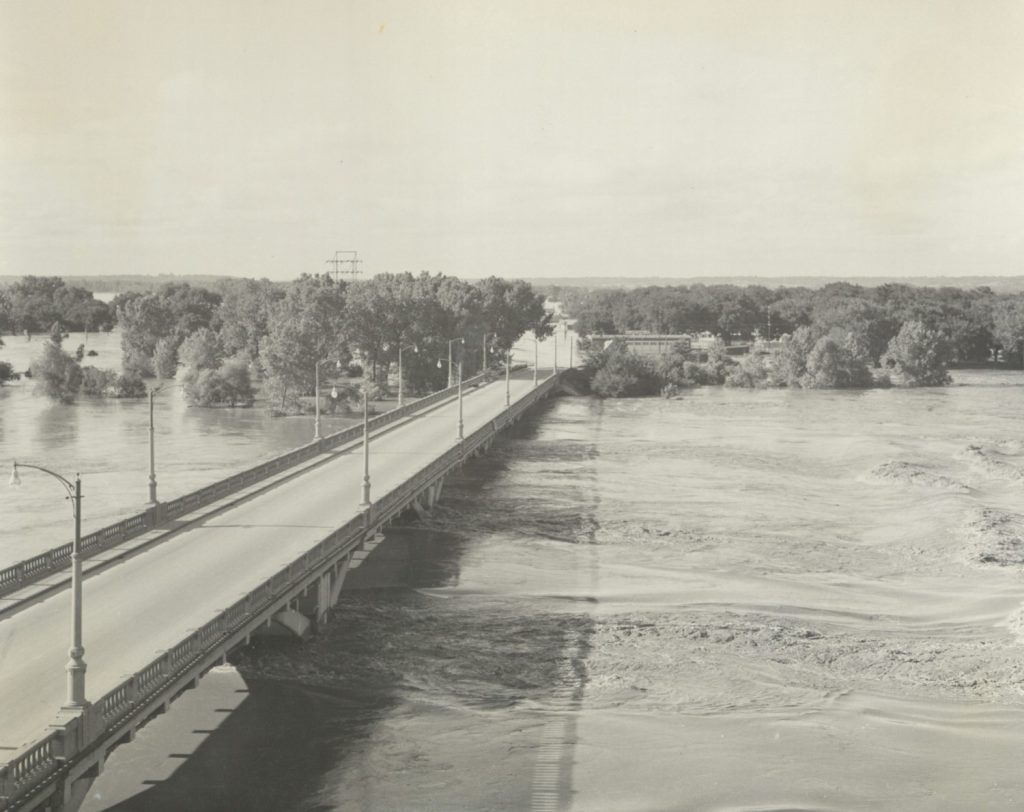 Black-and-white photograph of high water under a bridge.