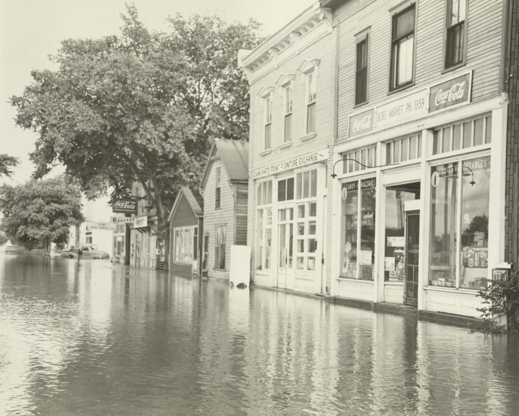 Black-and-white photograph of a row of buildings along a street flooded with several inches of water.