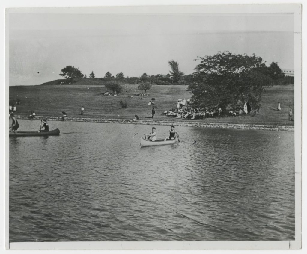 Black-and-white photograph of two pairs rows in canoes, with spectators on the grassy hill beyond.