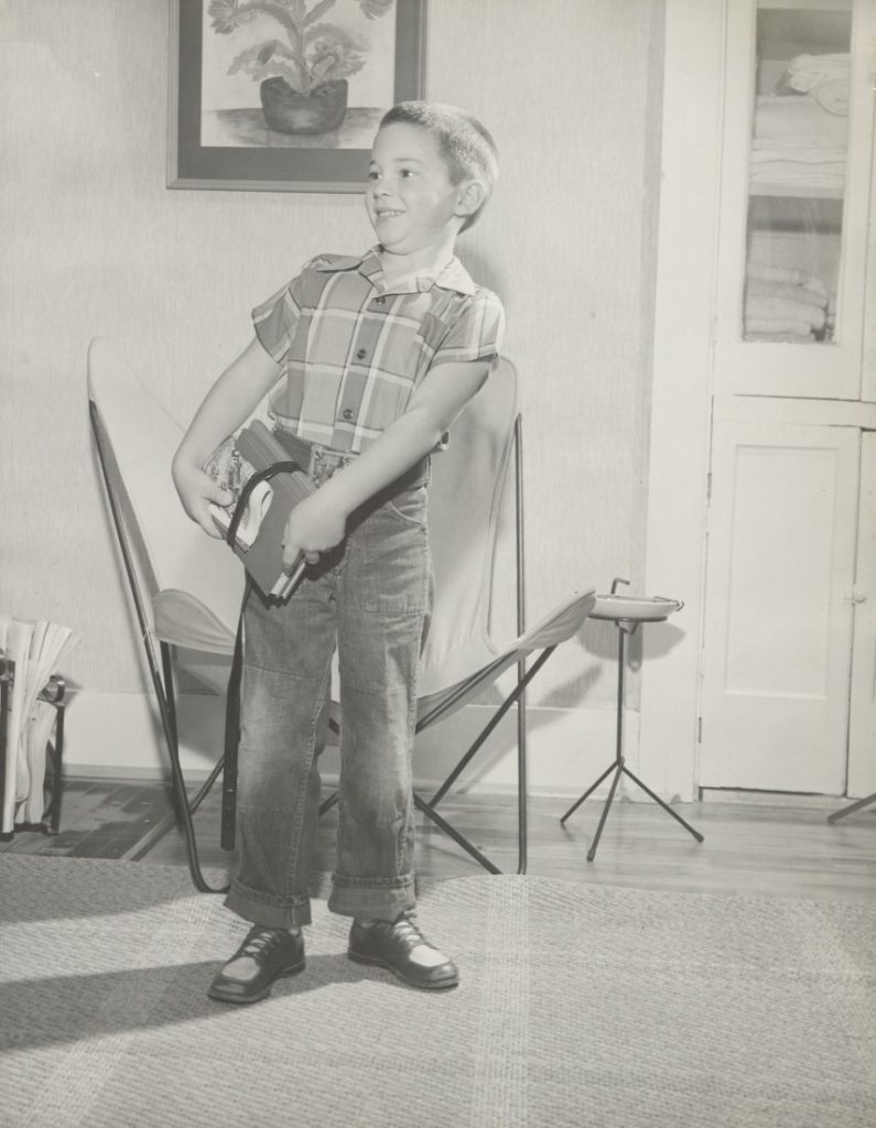 Black-and-white photograph of a young boy in long pants and a plaid short-sleeve shirt, standing at home and holding a couple of books at his hip.