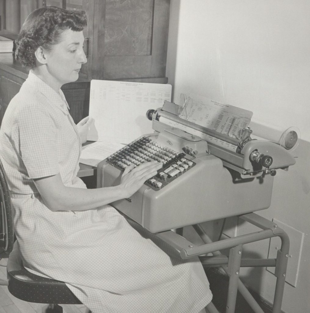 Black-and-white photograph of a woman sitting at a typewriter with a piece of paper in her hand.