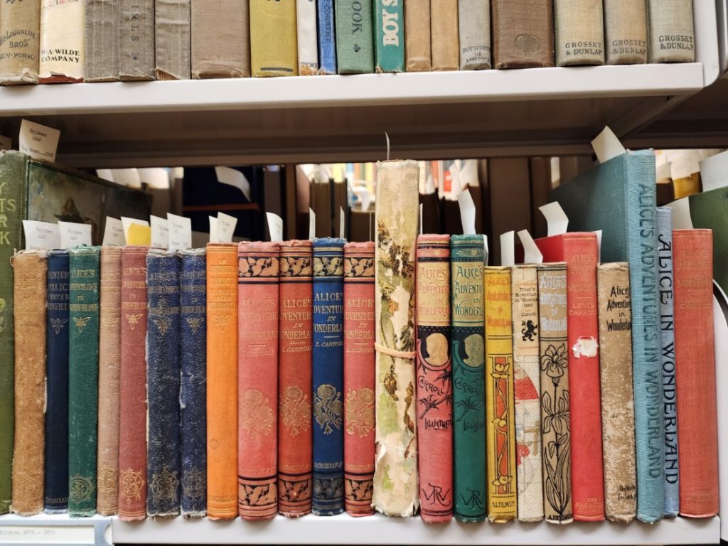 Color photograph of books - different editions of Alice's Adventures in Wonderland - lined up horizontally on a shelf.