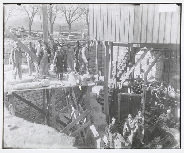 Black-and-white photograph of men standing with large machinery in an open-air underground area lined with bricks.