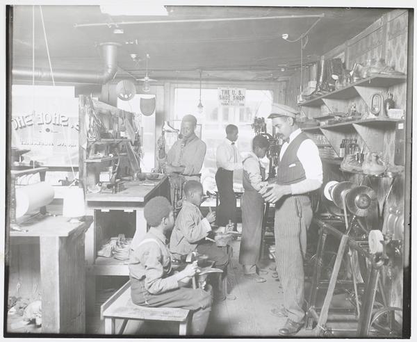 Black-and-white photograph of a crowded shop. Six boys and young men work on shoes: four stand and two sit on a bench.