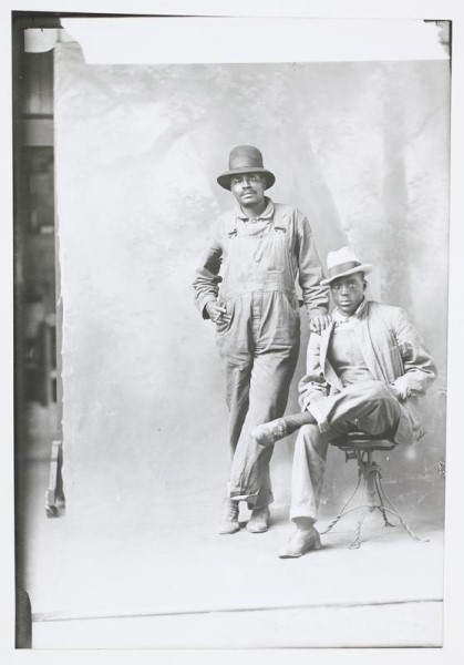 Blac-and-white photograph of two African American men in hats, long-sleeve shirts, and overalls. One is standing and one is sitting; they are in a studio in front of a backdrop. 