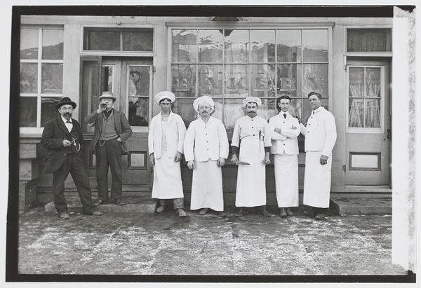 Black-and-white photograph of seven men standing in a row in front of a building. Five are dressed in chef's whites, and three are wearing hats.