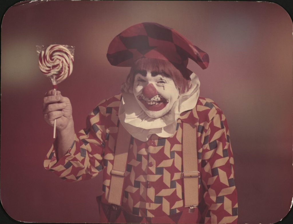Color photograph a smiling clown who is holding a large lollipop.