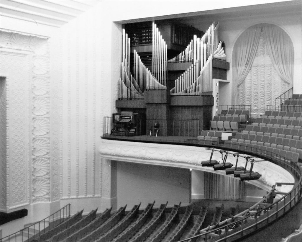 Black-and-white photograph of a pipe organ in the corner of a balcony in an auditorium.