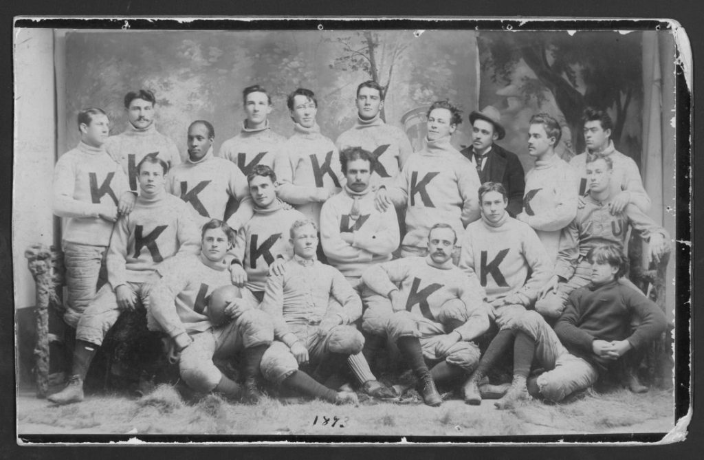 Black-and-white photograph of a group of young men; all are wearing light turtleneck sweaters with a large letter "K" on the front.