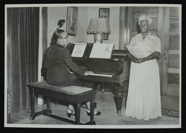 Black-and-white photograph of two African American women. One is playing the piano and one is singing.