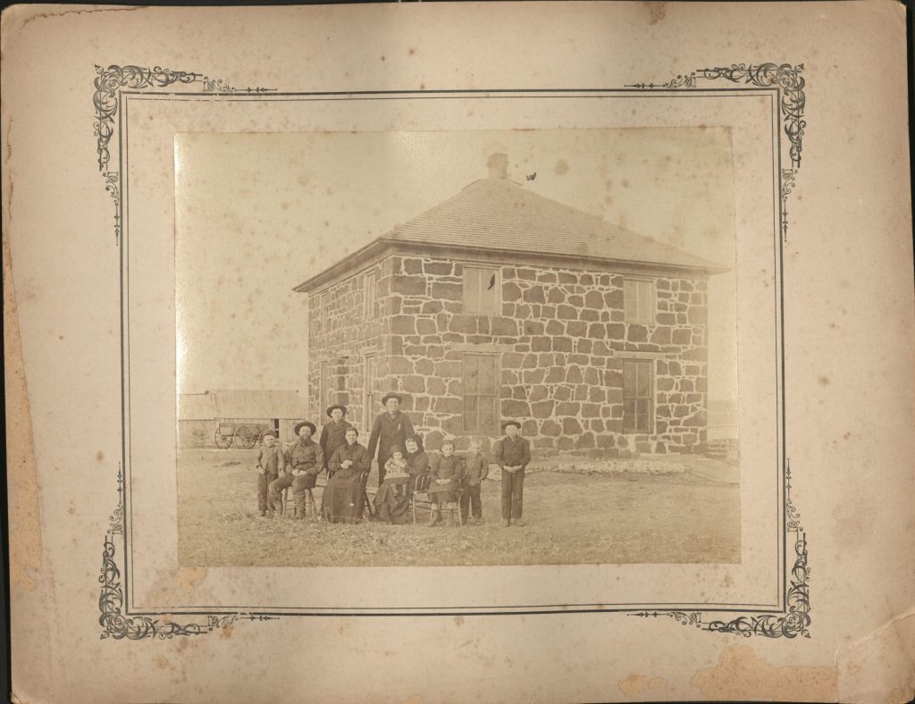 Sepia-toned photograph of a large inter-generational family in front of a two-story stone house.